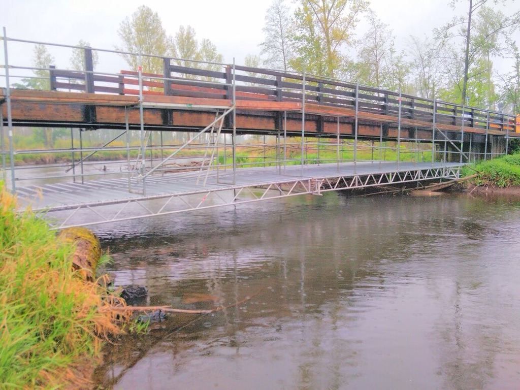 Behelfssteg aus Gerüstbauteilen führt über ein Gewässer, daneben eine hölzerne Brücke im Nebel